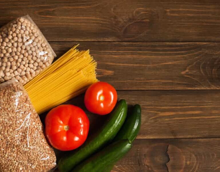 Dry beans, spaghetti pasta, and a grain in plastic bags are laid out on a wooden surface next to two tomatoes and two cucumbers. The food items are arranged in the lower left corner of the image.