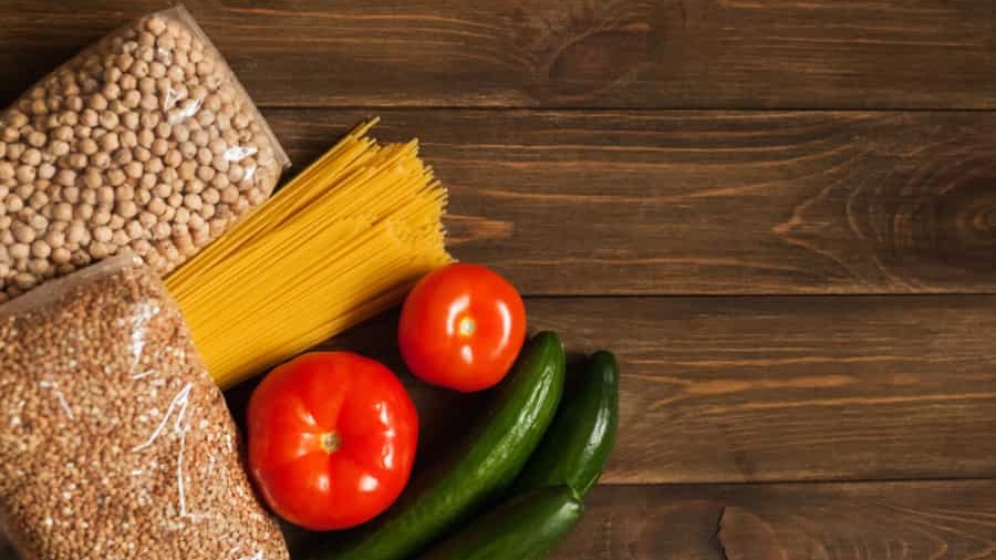 Dry beans, spaghetti pasta, and a grain in plastic bags are laid out on a wooden surface next to two tomatoes and two cucumbers. The food items are arranged in the lower left corner of the image.