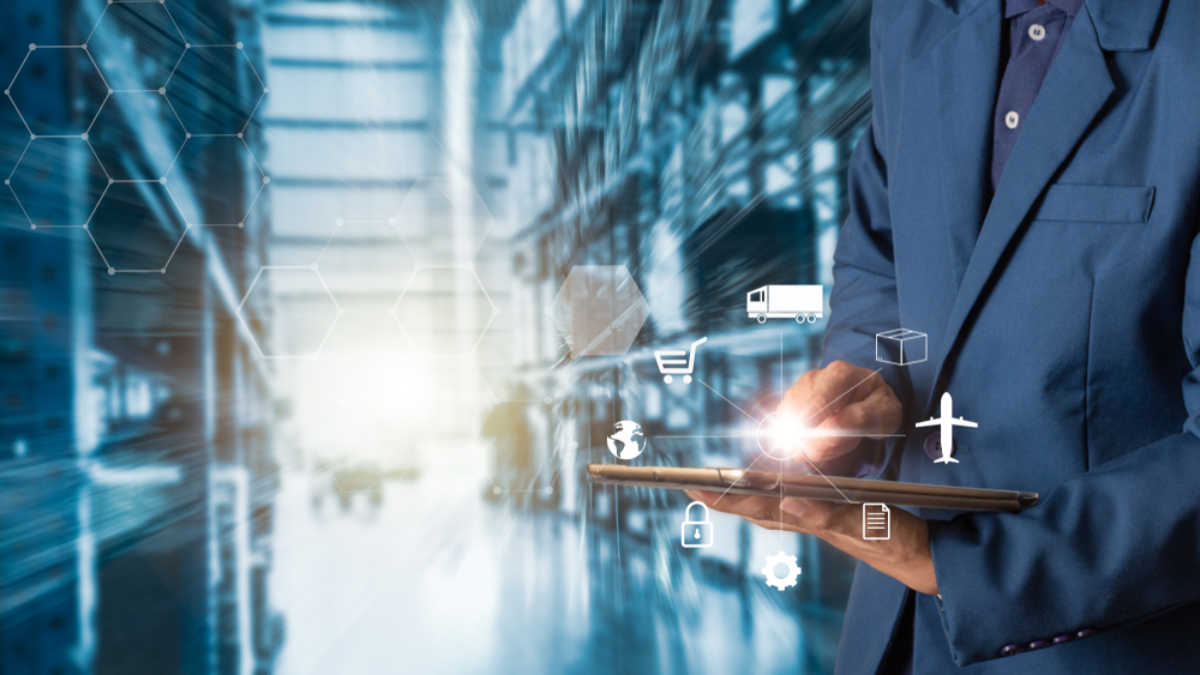 A person in a blue suit uses a tablet with virtual logistics and supply chain icons, such as a truck, airplane, and package, overlaid on an industrial warehouse background.