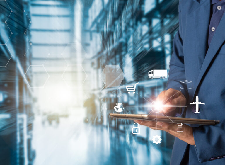 A person in a blue suit uses a tablet with virtual logistics and supply chain icons, such as a truck, airplane, and package, overlaid on an industrial warehouse background.