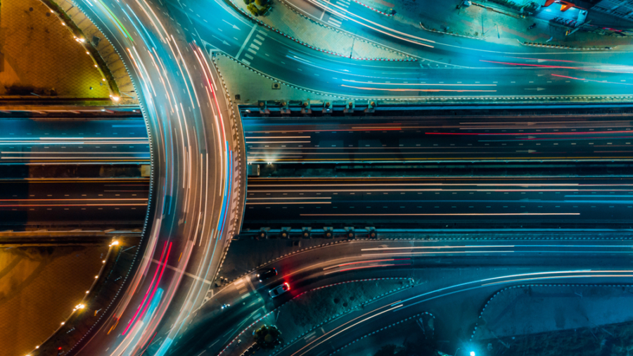 Aerial view of a brightly lit highway interchange at night, with streaks of red and blue lights from moving vehicles creating dynamic, colorful lines across multiple lanes and overpasses.