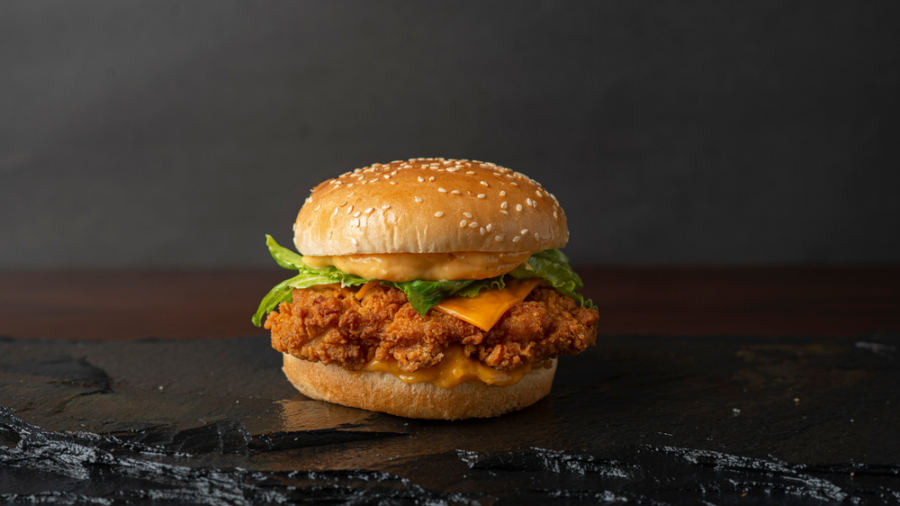 A crispy fried chicken sandwich with lettuce, cheddar cheese, and sauce in a sesame seed bun, placed on a dark slate surface with a black background.