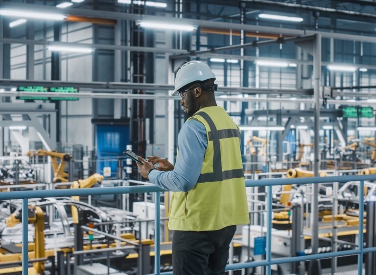 A worker wearing a hard hat and high-visibility vest uses a tablet while standing on a platform overlooking an automated factory floor with robotic arms and industrial equipment.
