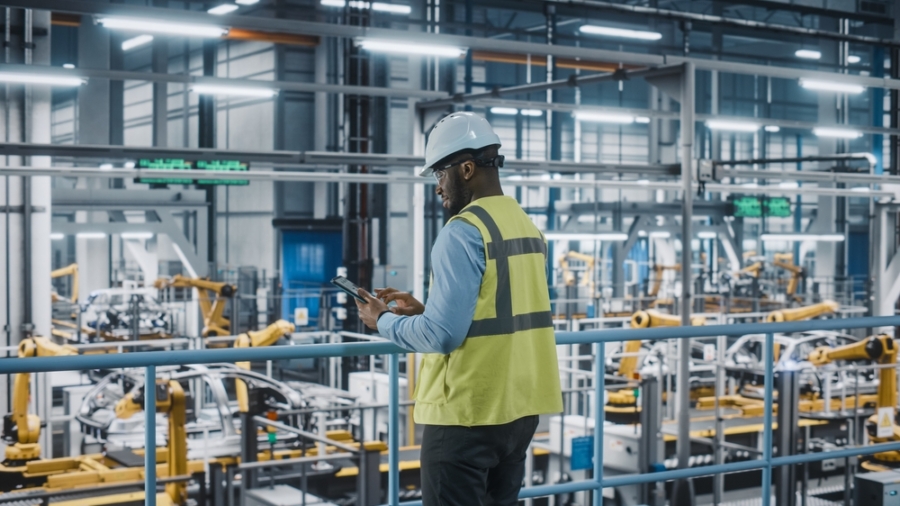A worker wearing a hard hat and high-visibility vest uses a tablet while standing on a platform overlooking an automated factory floor with robotic arms and industrial equipment.