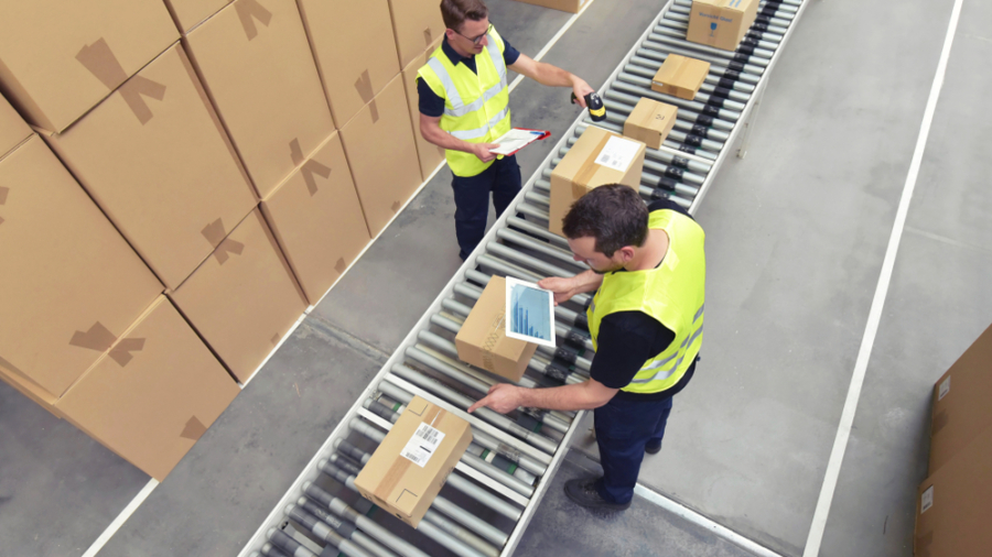 Two warehouse workers in yellow safety vests scan and check boxes on a conveyor belt; large stacks of cardboard boxes are visible nearby on the warehouse floor.