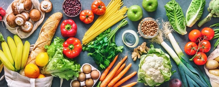 A variety of fresh groceries, including vegetables, fruits, eggs, pasta, bread, beans, oil, and milk, are arranged on a table. The colorful assortment features tomatoes, carrots, lettuce, apples, mushrooms, and potatoes.