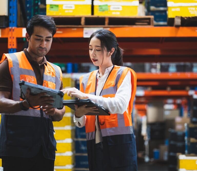 Two warehouse workers wearing orange safety vests review labor management details on a digital tablet, standing among shelves filled with boxes and containers in a storage facility.