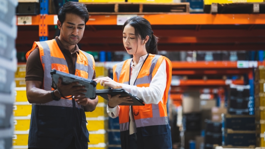 Two warehouse workers wearing orange safety vests review labor management details on a digital tablet, standing among shelves filled with boxes and containers in a storage facility.