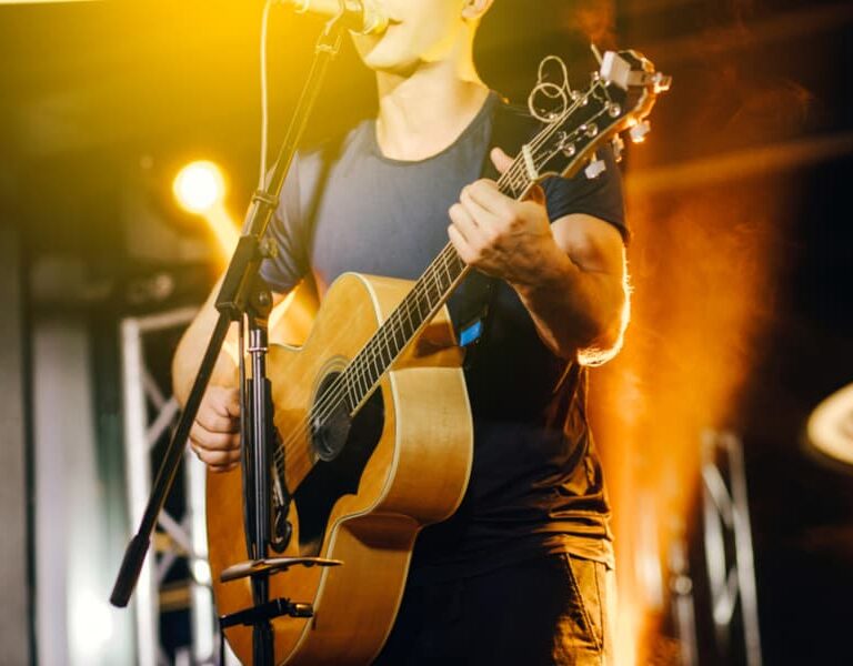 A musician in a dark t-shirt plays an acoustic guitar and sings into a microphone on a brightly lit stage with orange and yellow lights in the background.