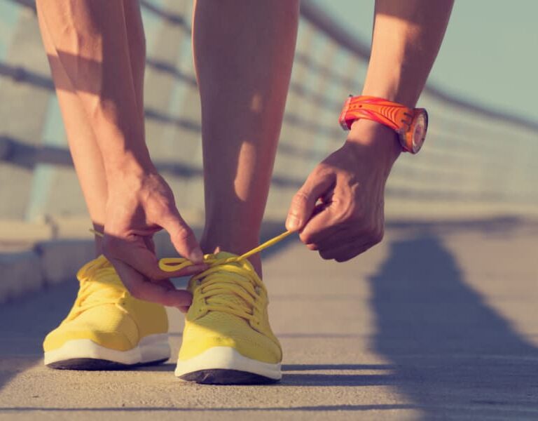 A person wearing a red wristwatch bends down to tie the laces of bright yellow running shoes on a sunlit outdoor path with a railing.