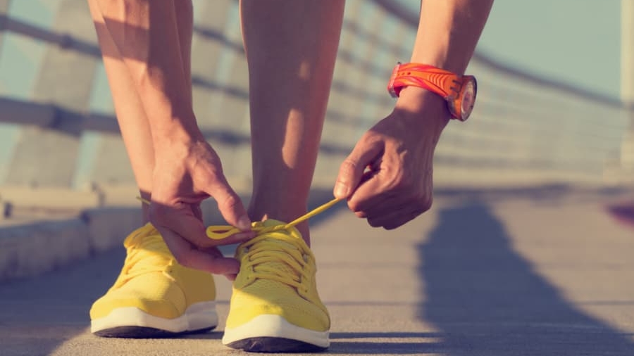 A person wearing a red wristwatch bends down to tie the laces of bright yellow running shoes on a sunlit outdoor path with a railing.