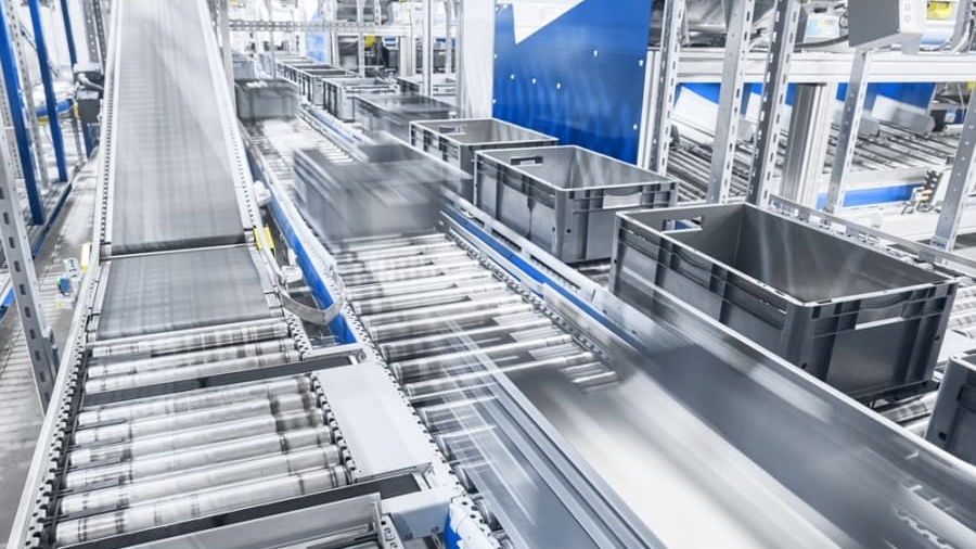 A modern automated conveyor system efficiently transports black plastic containers through a bright, industrial facility, with machinery and metal rollers moving at high speed to automate material handling.