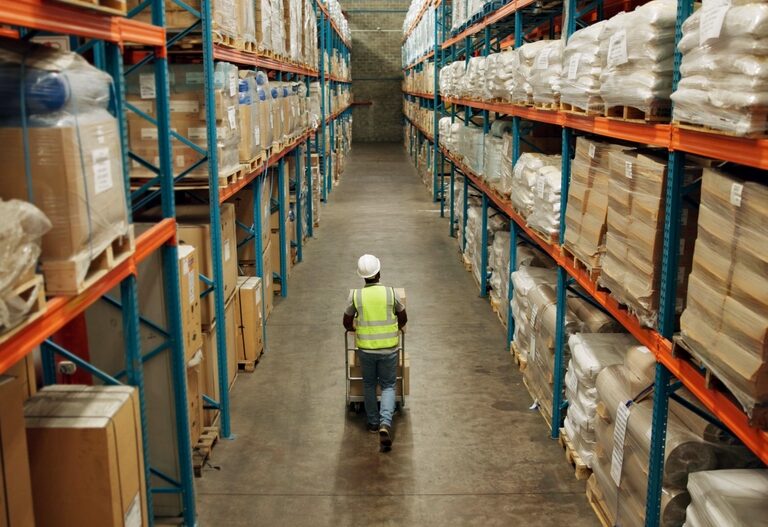 A warehouse worker wearing a reflective vest and hard hat pushes a cart down an aisle lined with tall shelves filled with boxes and wrapped pallets.