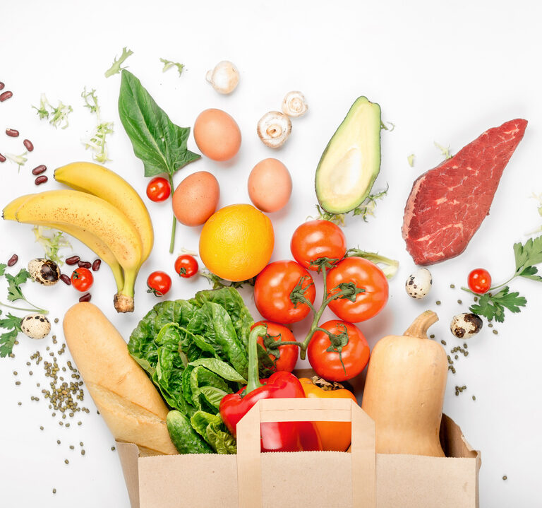 A paper bag overflows with fresh food—tomatoes, lettuce, bananas, bell pepper, baguette, eggs, and more—showcasing the importance of supply chain traceability on a white background.