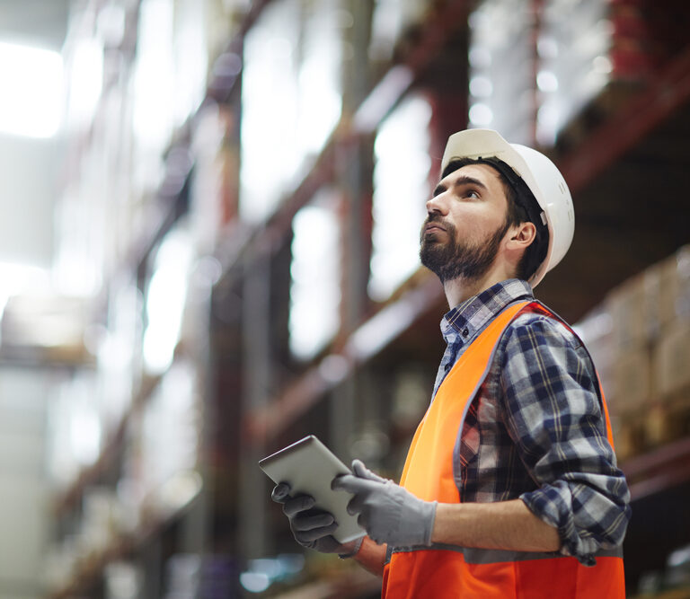 A warehouse worker in a hard hat and orange safety vest holds a tablet while inspecting shelves, ensuring that master planning keeps the large, well-lit storage facility organized and efficient.