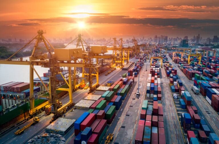 A busy shipping port at sunset, with large cranes, colorful cargo containers, and city skyline in the background. The sky is orange and the sun is low on the horizon.