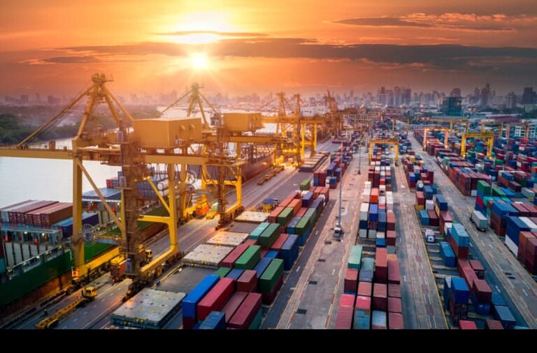 Shipping containers are stacked in rows at a busy port with large yellow cranes, as the sun sets over a city skyline in the background. The scene shows bustling industrial activity near a river.