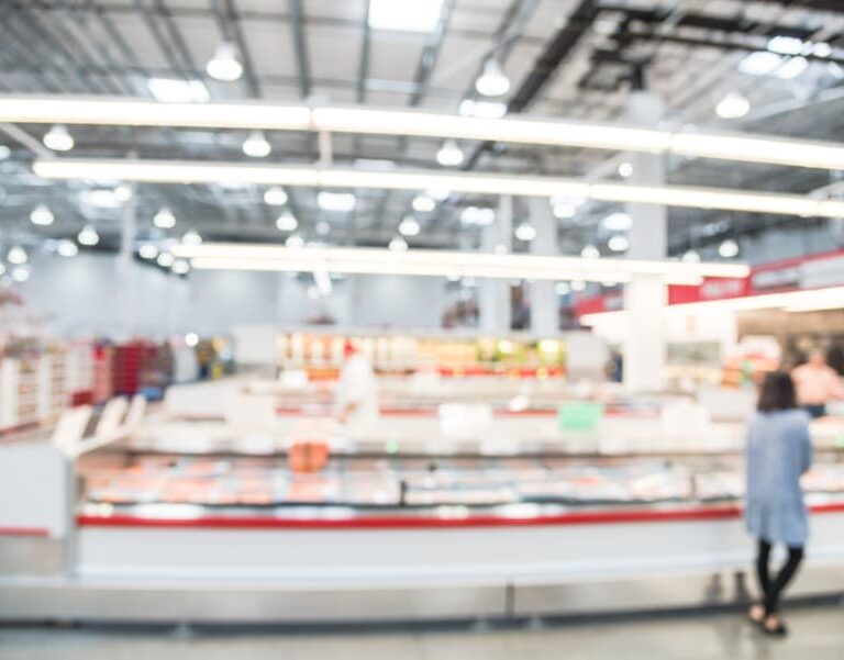 A blurry image of a person standing in a brightly lit supermarket or warehouse store, with shelves and display cases in the background. The scene is out of focus, making details difficult to distinguish.