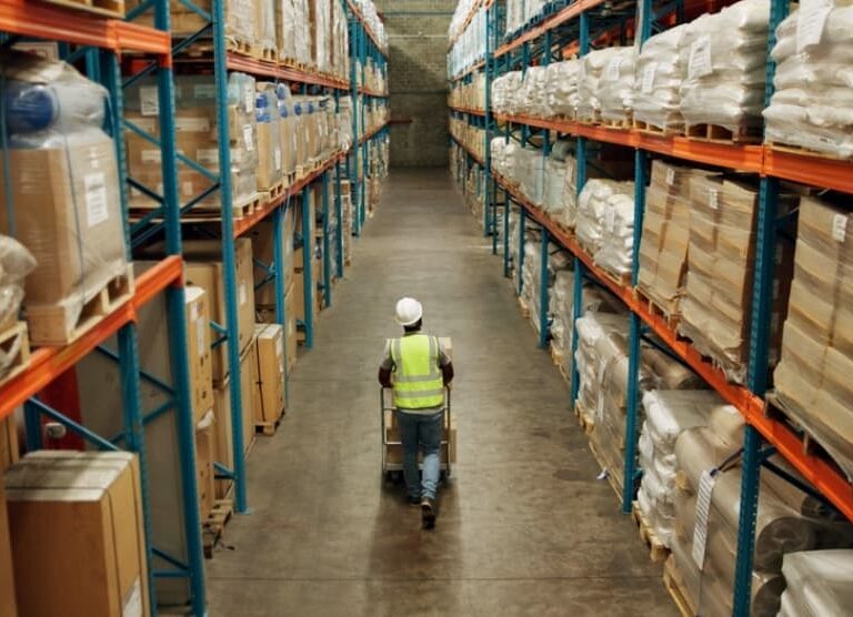 A worker in a high-visibility vest and hard hat pushes a cart down a wide aisle between tall warehouse shelves filled with boxes and packaged goods at Blue Ridge.