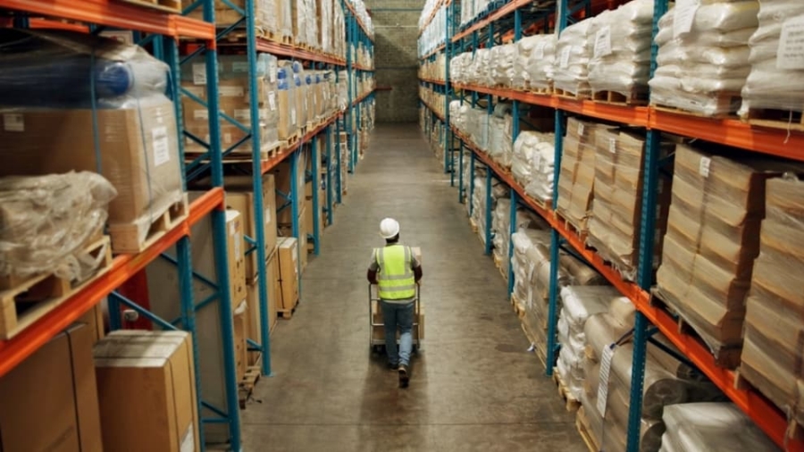 A worker in a high-visibility vest and hard hat pushes a cart down a wide aisle between tall warehouse shelves filled with boxes and packaged goods at Blue Ridge.