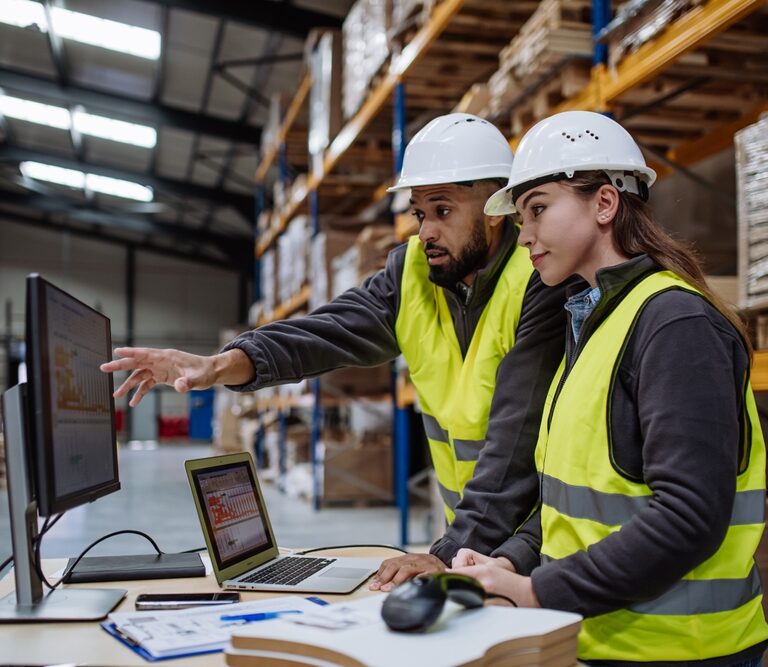 Two workers wearing hard hats and yellow safety vests look at a computer screen in a warehouse, focusing on order accuracy as one points at the monitor while the other observes. Shelves of boxes and pallets fill the background.