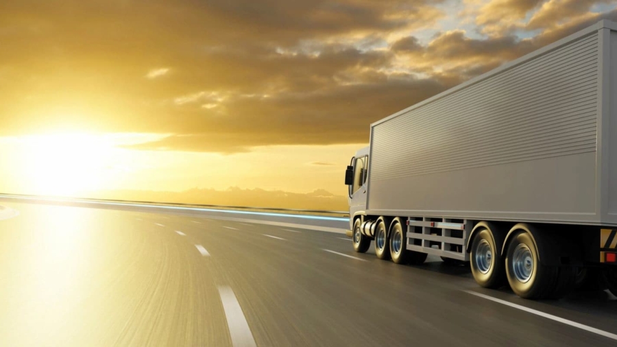 A large white truck drives on a highway toward the sunset under a cloudy sky, with sunlight reflecting off the road—undeterred by changing tariffs that shape its journey.