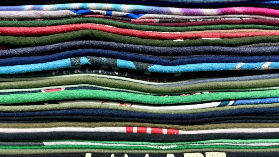 A close-up view of a neatly stacked pile of colorful folded t-shirts, arranged in horizontal layers showing various patterns and textures, highlights the variety available despite recent tariff changes.