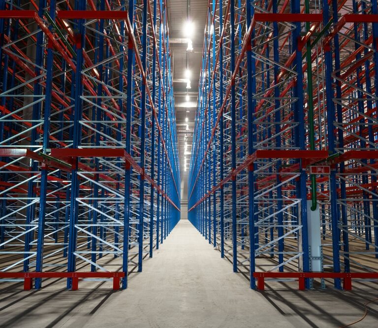 Rows of tall, empty metal storage racks in a brightly lit warehouse, forming a symmetrical, industrial corridor—standing ready for inventory as tariff changes impact the flow of goods.