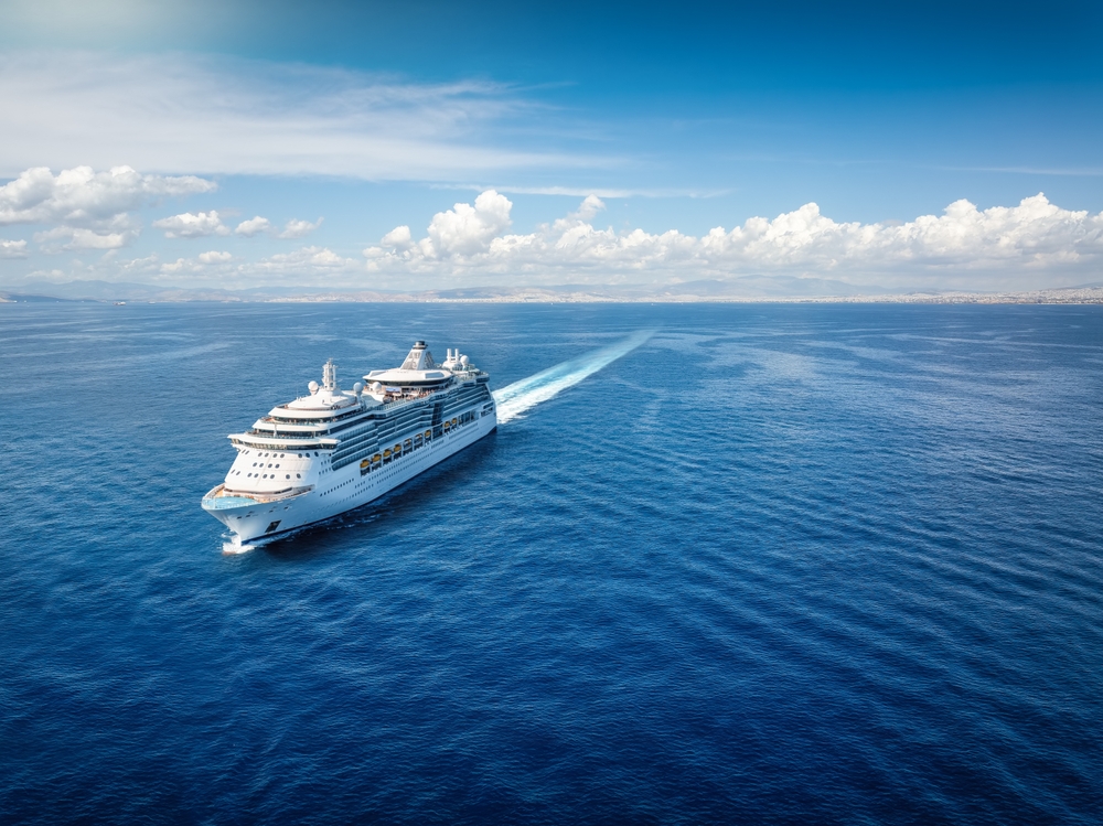 A large white cruise ship, known for its all-inclusive tariff, sails across a calm, deep blue sea under a bright blue sky with scattered clouds, leaving a white trail behind. Land is visible on the distant horizon.