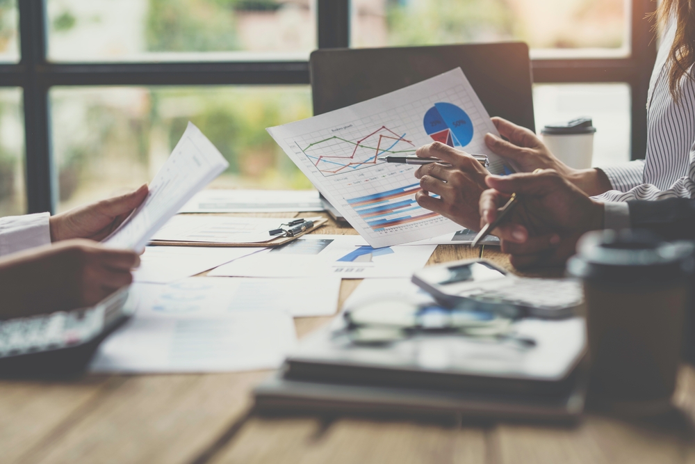 People sitting at a table review financial documents and charts with graphs and data in a bright office setting. Laptops, papers, and coffee cups are visible on the wooden table.