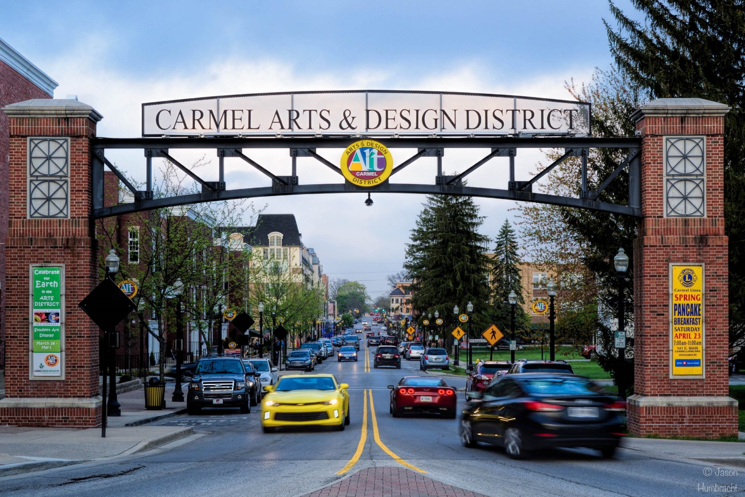 A street scene under the Carmel Arts & Design District archway, with cars driving on the road, including a bright yellow car in the foreground, and shops and trees lining both sides.