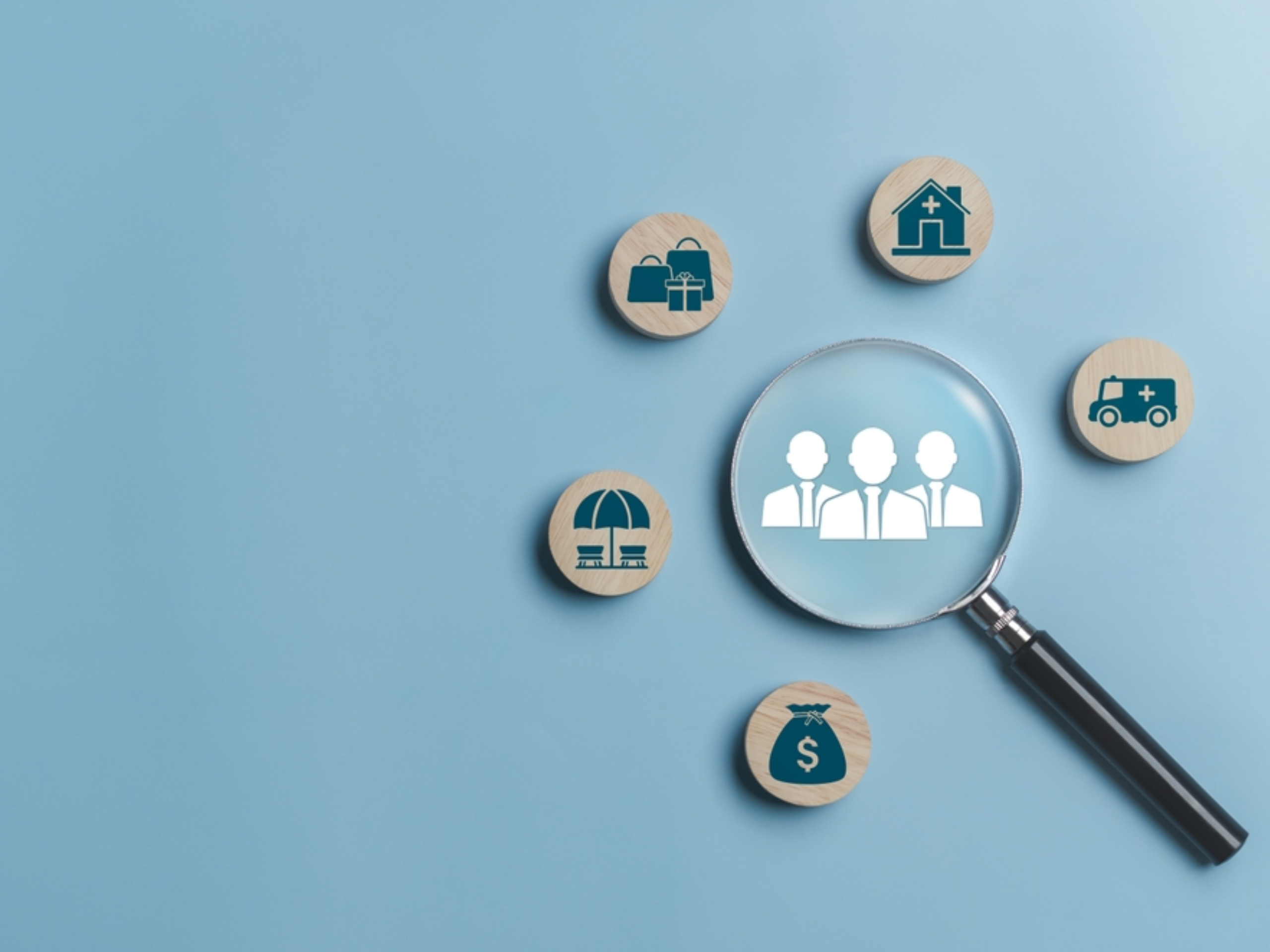 A magnifying glass highlights a wooden token with three people icons, surrounded by tokens showing a house, ambulance, shopping bags, umbrella, and a money bag on a blue background.