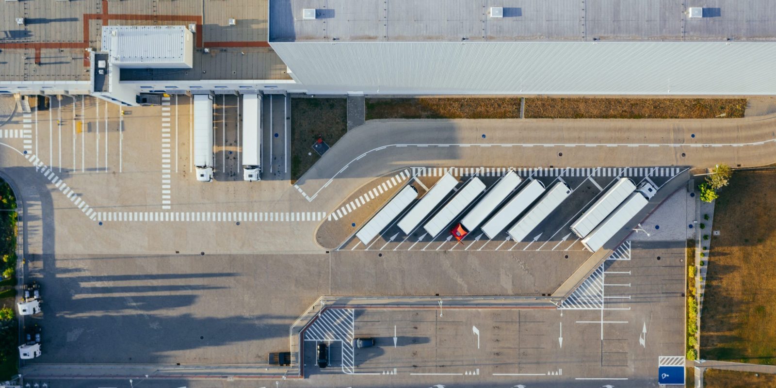 Aerial view of a large warehouse with multiple loading docks, several semi-trailers parked in a row, and a parking lot with cars in the lower section of the image.