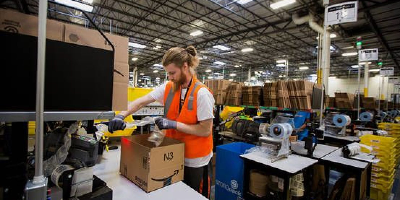 A warehouse worker in an orange safety vest packs items into a cardboard box at a workstation in a large, brightly lit industrial facility filled with equipment and stacks of boxes.