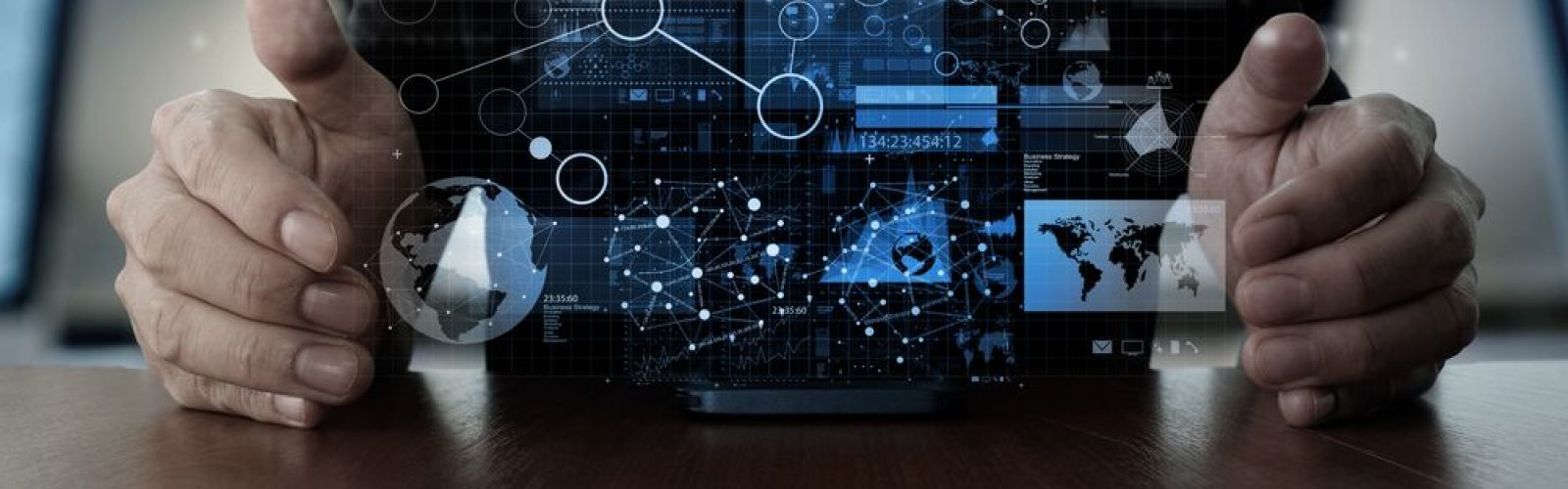 A person’s hands are hovering around a digital interface with floating futuristic graphics, including charts, maps, and network connections, projected above a desk. The scene suggests technology, data analysis, or innovation.