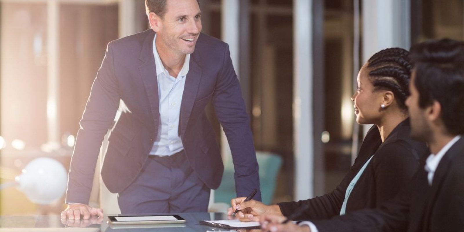 A man in a blue suit stands and smiles while speaking to two colleagues, a woman and a man, seated at a conference table with notebooks and a laptop in a modern office setting.