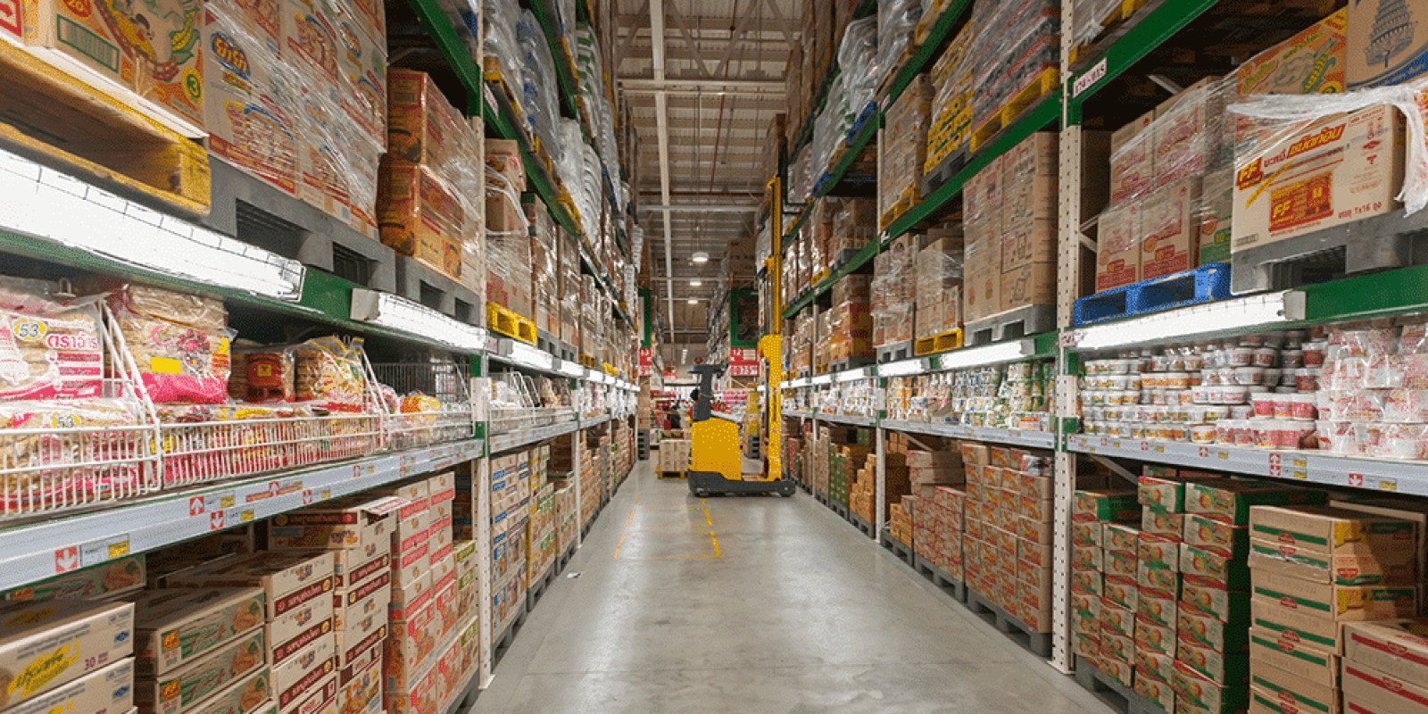 A wide aisle in a warehouse-style grocery store features tall shelves stacked with packaged food products, highlighting the importance of 3PL selection for efficient logistics, with a yellow pallet jack parked at the end of the aisle.