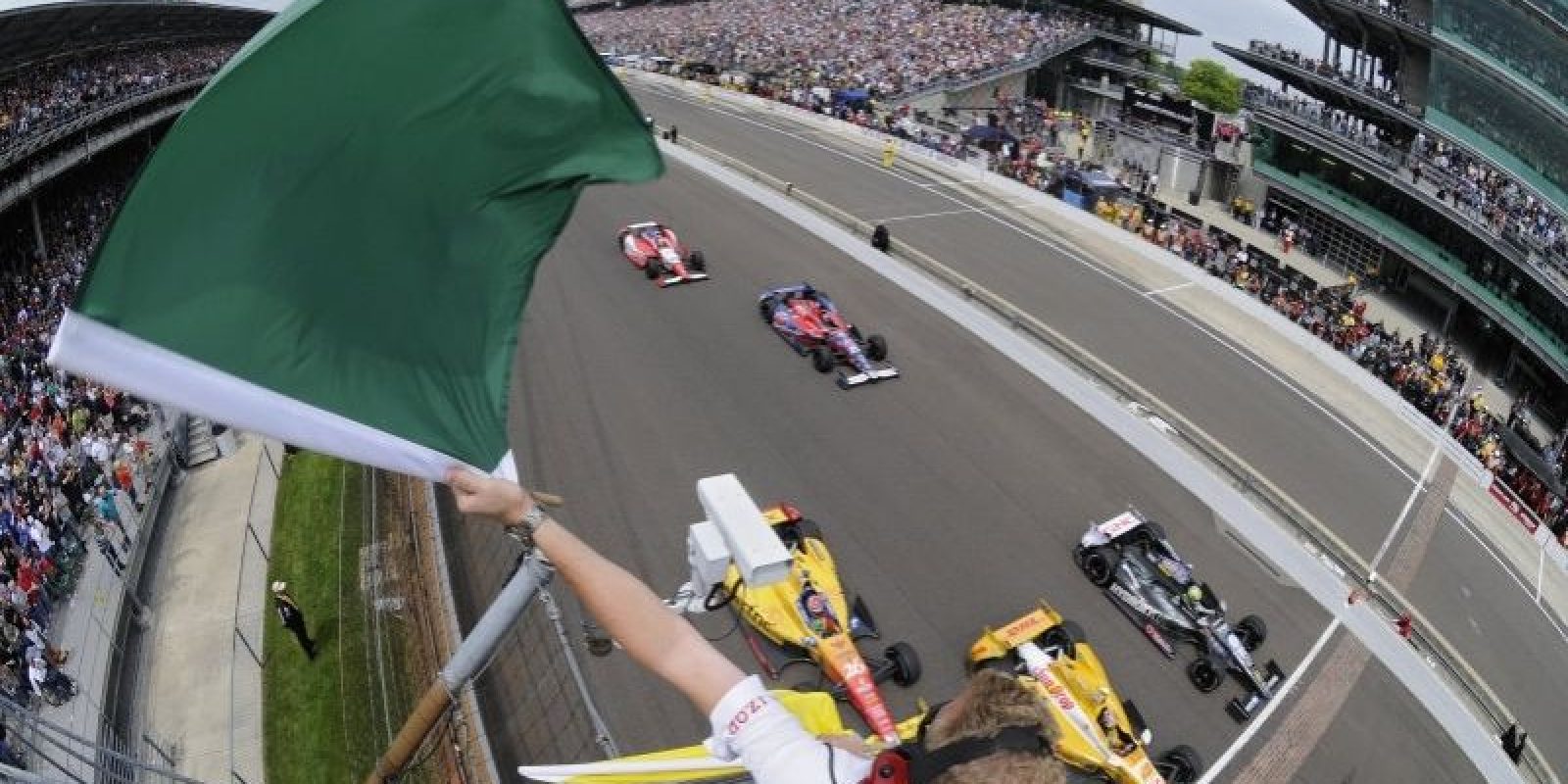 A race official waves a green flag to start an IndyCar race, with cars speeding down the track and a packed grandstand of spectators in the background.