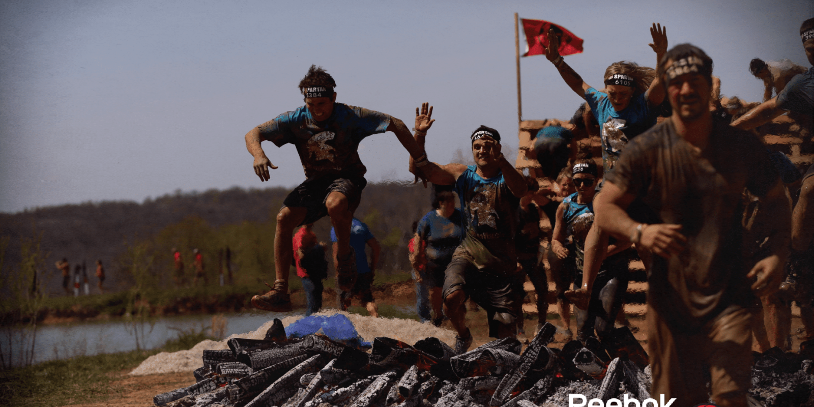 Group of people in athletic gear and headbands leap over burning logs during a muddy outdoor obstacle course race, with a Reebok Spartan Race logo in the bottom right corner.