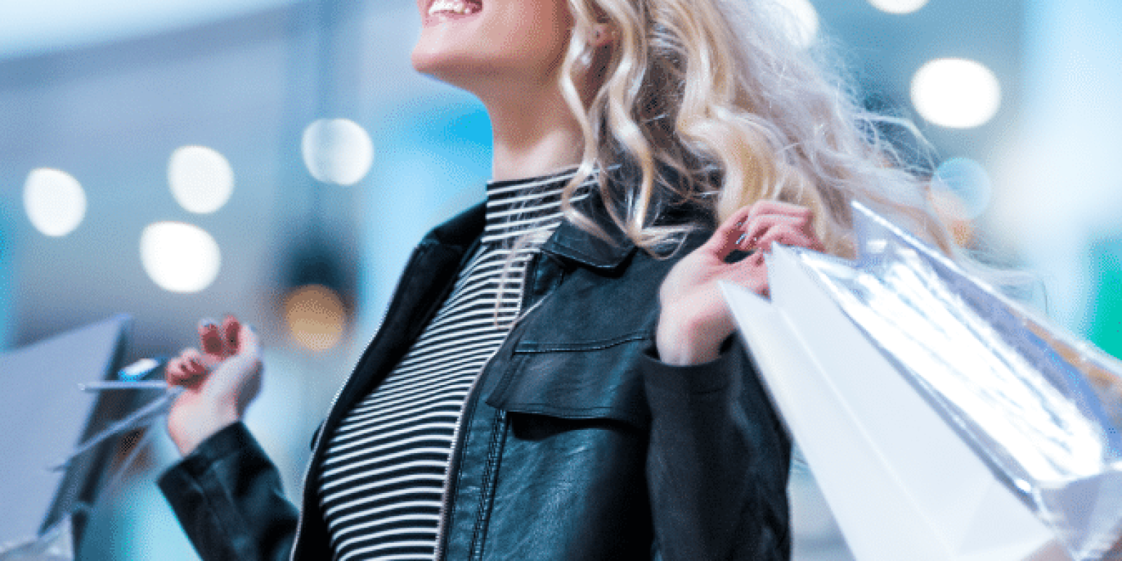 A smiling woman with long blonde hair wearing a striped top and black leather jacket holds several shopping bags in a brightly lit mall.