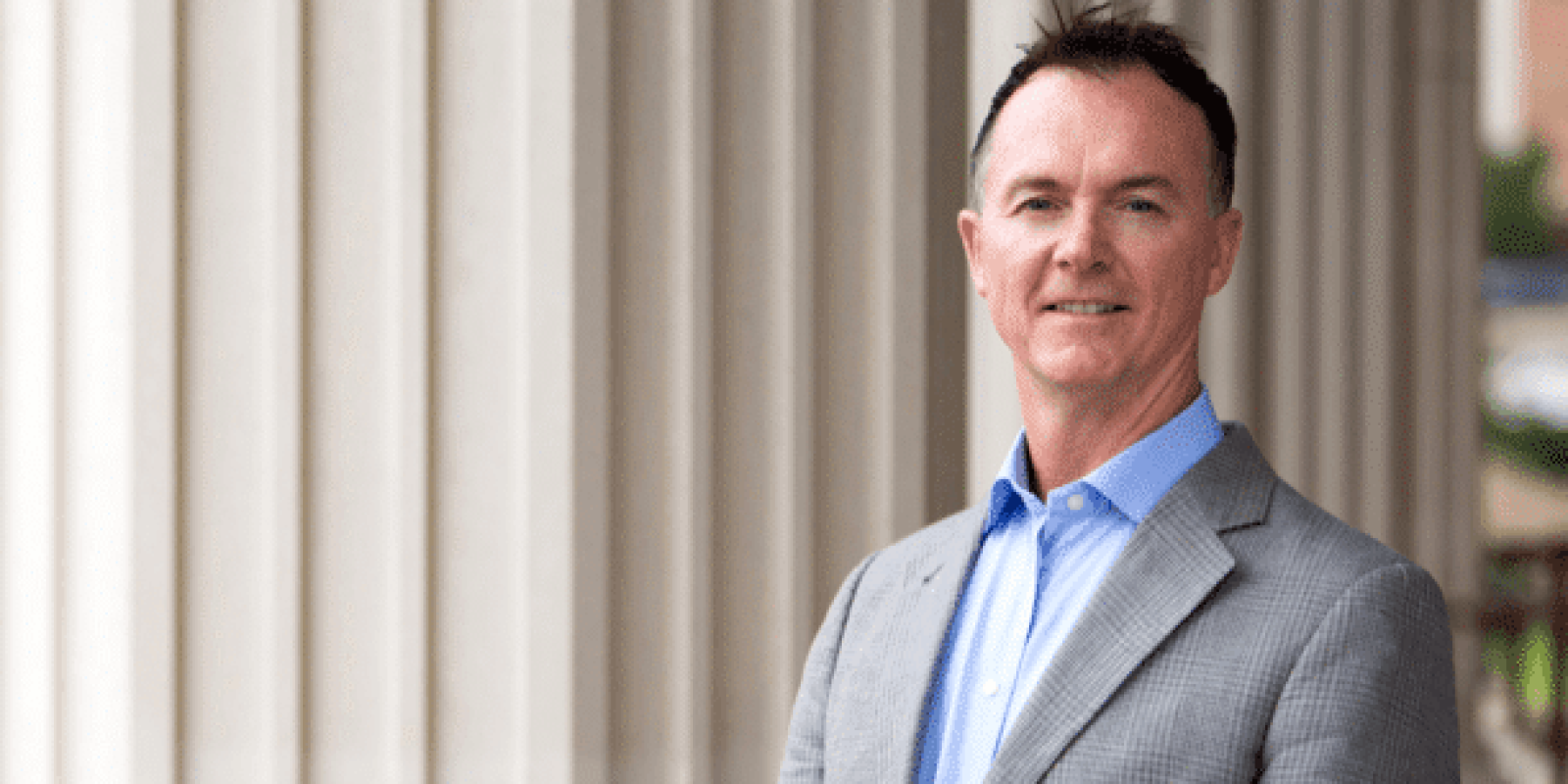 A man in a light gray suit and blue shirt stands outside in front of tall white columns, smiling at the camera.