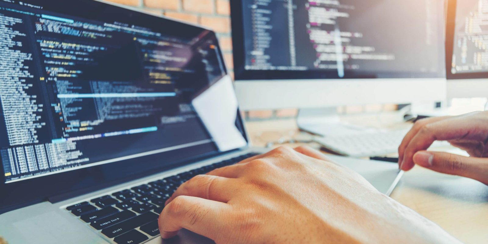 A person types on a laptop with code displayed on the screen. Two large monitors, also showing code, are on a desk in the background—a modern workspace ideal for tech roles like those at enVista Careers.