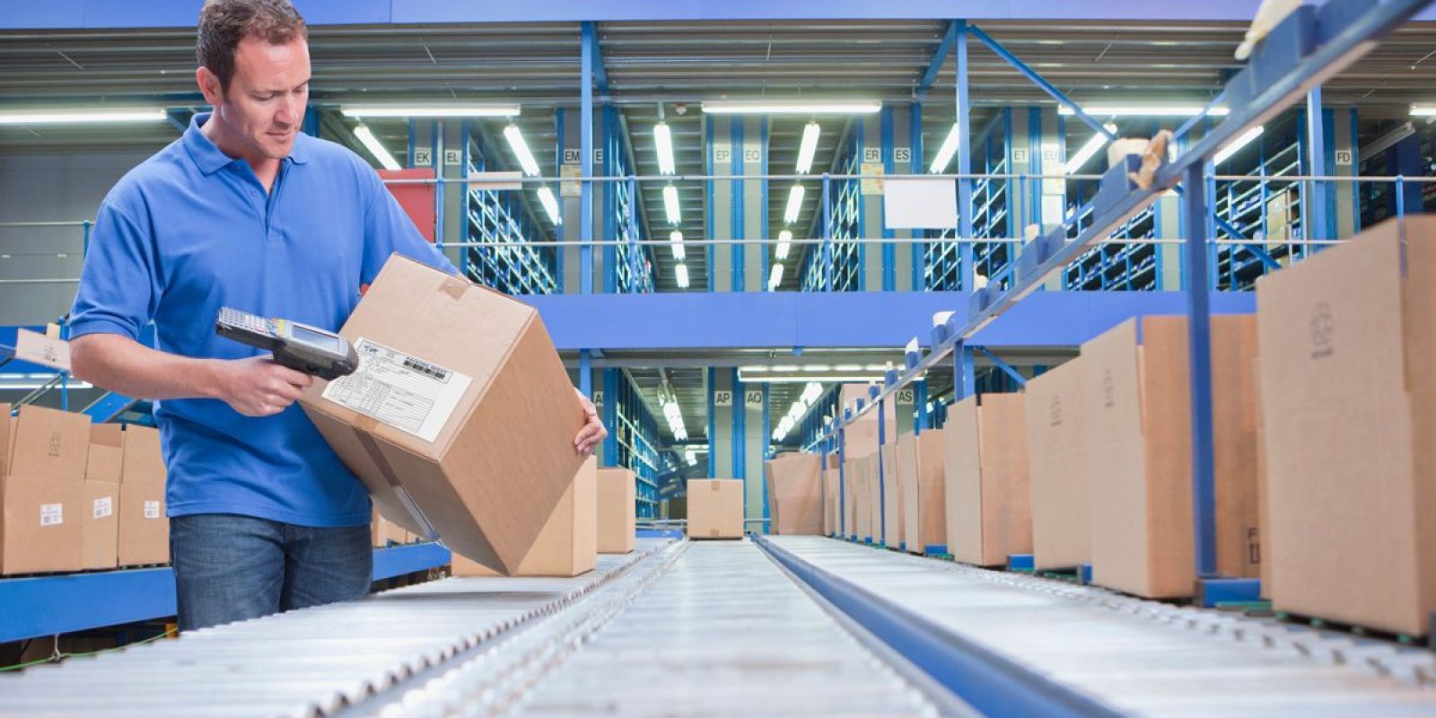 A man in a blue shirt scans a shipping label on a cardboard box in a warehouse, standing beside a conveyor belt with several other boxes—a scene streamlined by distributed order management under bright industrial lights.