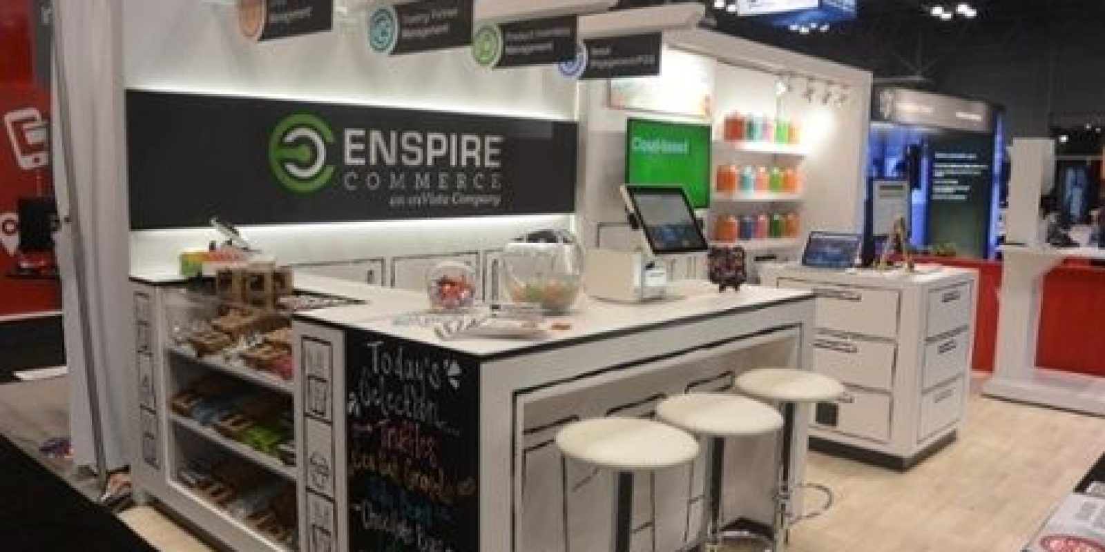A trade show booth for Enspire Commerce with a countertop, white stools, computer screens, and organized shelves of snacks. Bright signs and colorful jars are displayed in a well-lit exhibition space.