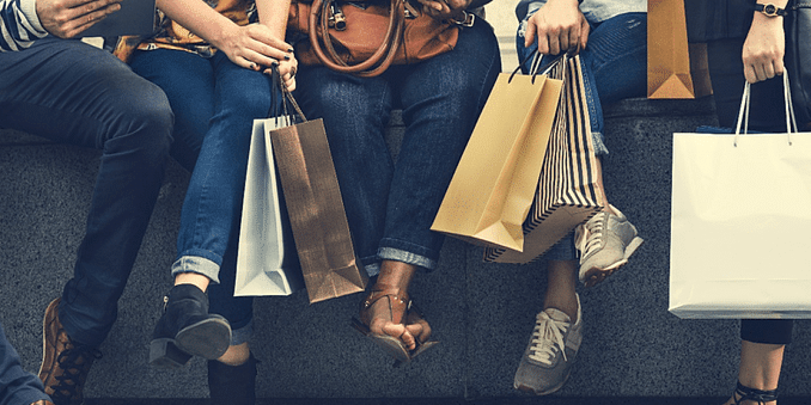 Four people sit on a bench holding various shopping bags. Only their legs, hands, and bags are visible. They wear casual clothing and different types of shoes, suggesting a shopping outing.