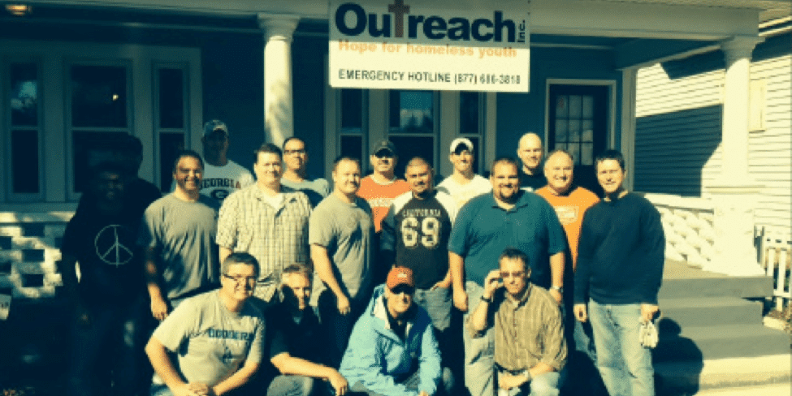 A group of men pose and smile in front of a white house with a sign reading “Outreach: Hope for Homeless Youth. Emergency Hotline (877) 614-3818.” The group stands on grass under bright sunlight.