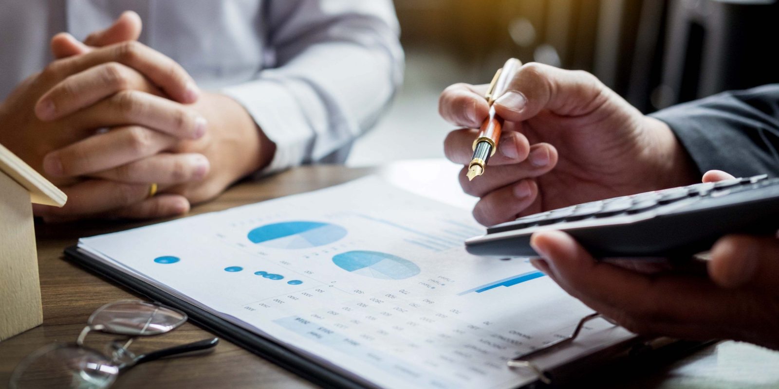 Two people sit at a desk; one holds a pen and calculator, the other has clasped hands. A clipboard with financial charts, glasses, and an enVista 365 pricing sheet are on the table, suggesting a discussion about finances or planning.