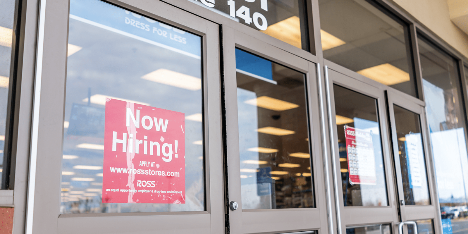 Glass doors of a retail store with a prominent red Now Hiring! sign displayed, reflecting the parking lot outside; store lights and aisles are visible through the glass.