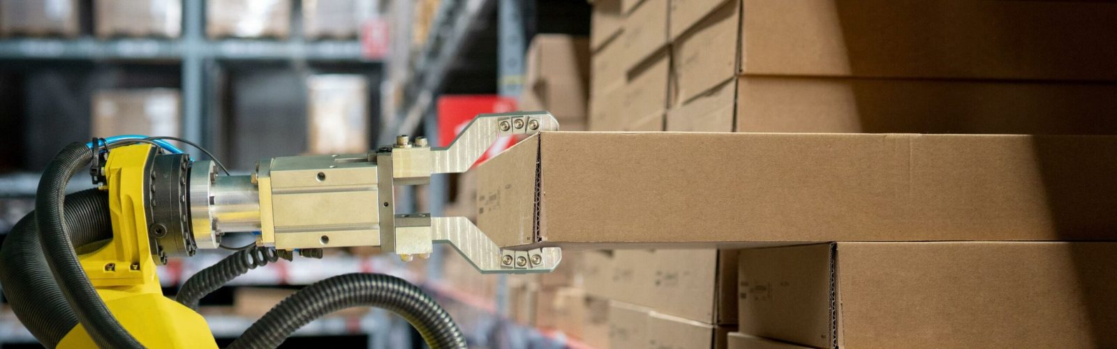 A robotic arm lifts a cardboard box from a shelf filled with similar boxes in a warehouse, illustrating automation in inventory management.