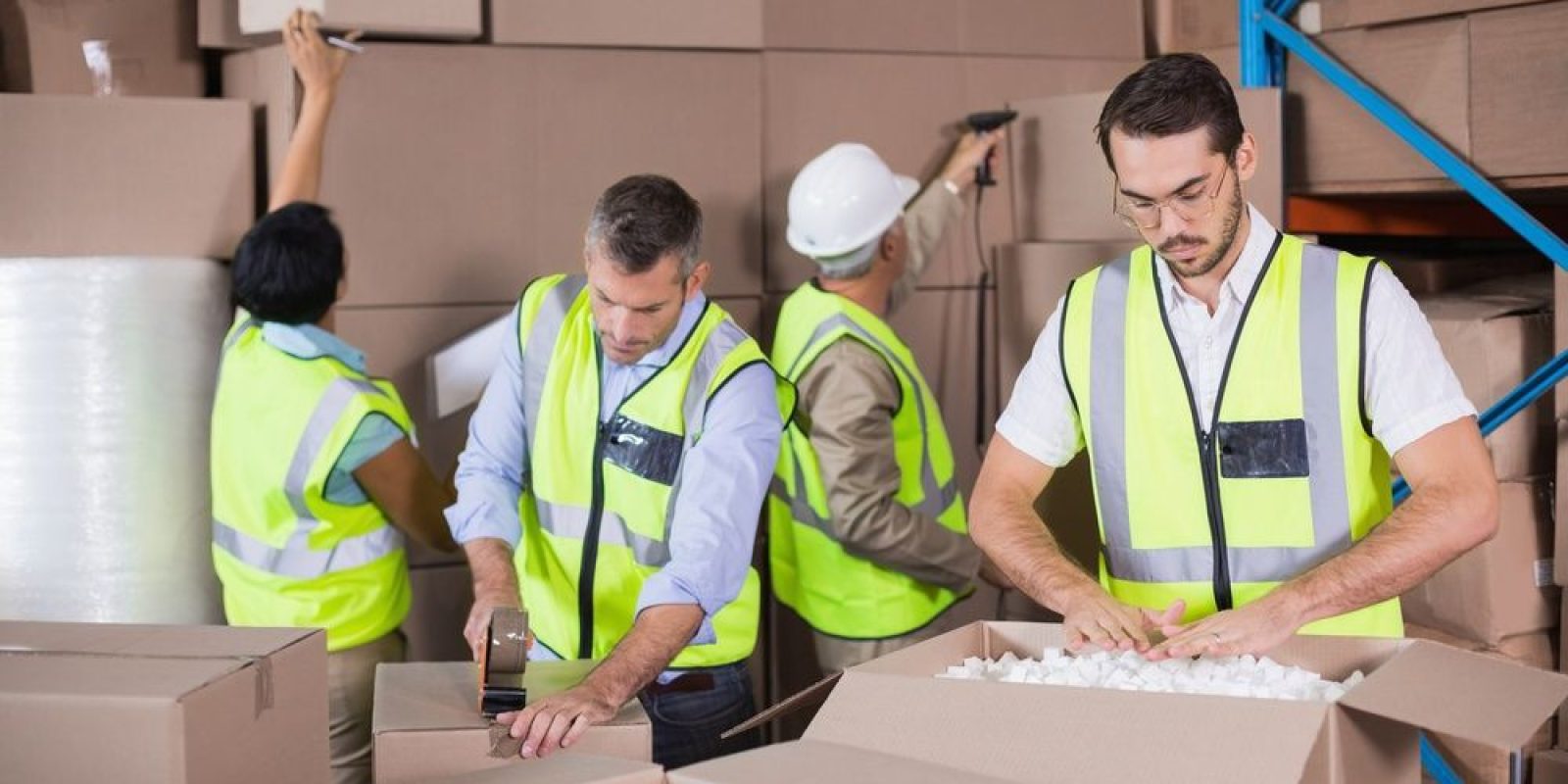 Four workers in safety vests organize and pack boxes in a warehouse. One person tapes a box, another fills a box with packing peanuts, and two others stack boxes on shelves.